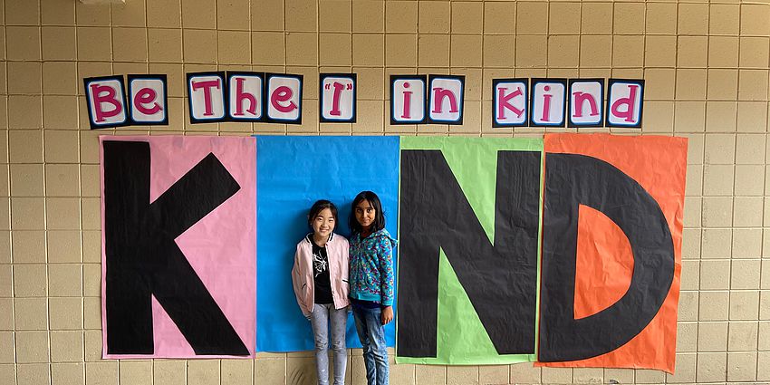 two students standing in front of a "Kind" poster