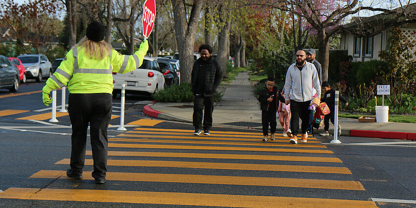 woman crossing guard holds a stop sign as a couple of children and adults crosse the street marked with wide yellow lines.