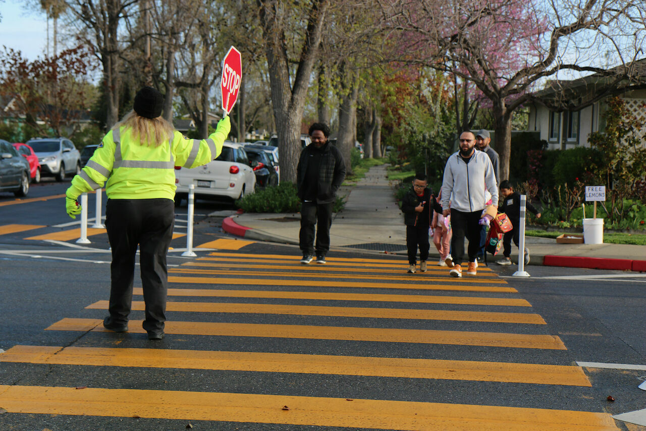 woman crossing guard holds a stop sign as a couple of children and adults crosse the street marked with wide yellow lines.